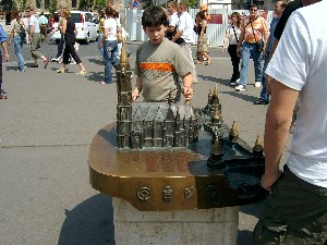 A boy with dark hair is looking at the model and touching one of the spires of the church. Other people are standing around looking at the model and in the background are groups of tourists.